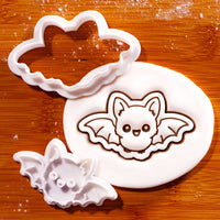 Flying Chibi Bat Mini Cookie Cutter and matching stamp set displayed on a wooden surface beside rolled white fondant embossed with a cute flying bat design.