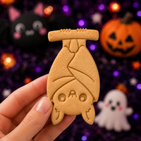 Embossed hanging chibi bat cookie held by hand against a spooky Halloween background with pumpkins and ghosts