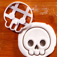 Cute Skull front view cookie cutter beside a detailed skull impression in white fondant on a wooden baking surface