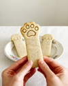 Hand holding a cookie shaped like a cat paw print with two more cat paw cookies on a decorative plate in the background.